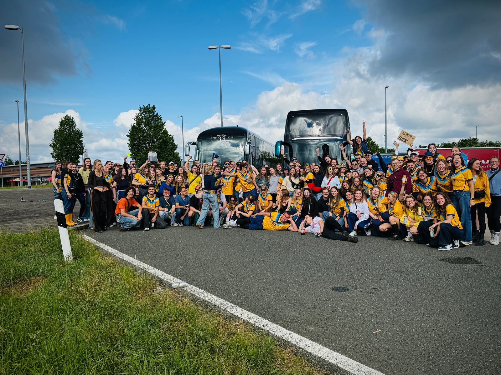 Large student group in yellow and blue jerseys in front of two coaches at a motorway stop